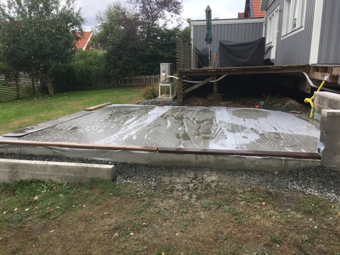 Nyligen gjuten och glättad betongplatta vid husgrund med gräsmatta i bakgrunden.