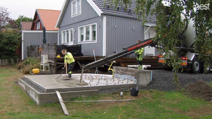 Nyglättad betongplatta med arbetare och betongbil i bakgrunden vid en husgrund.