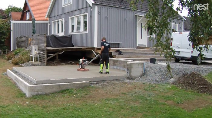 Nyglättad betongplatta med arbetare och glätningsmaskin framför hus under renovering.