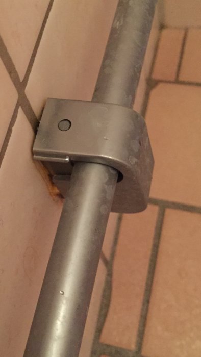 Brun/orange missfärgning bakom rörfäste på kakelvägg som rinner ner längs fogen, möjlig vattenskada.