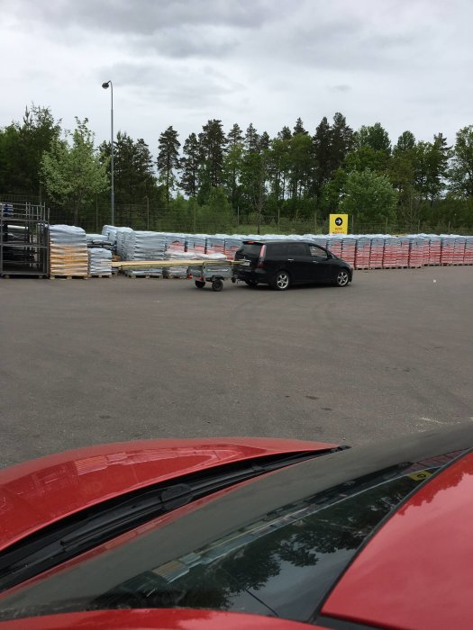 Svart bil parkerad på ett öppet område med släpvagn kopplad, vid byggmaterial och skogsrabatt i bakgrunden.