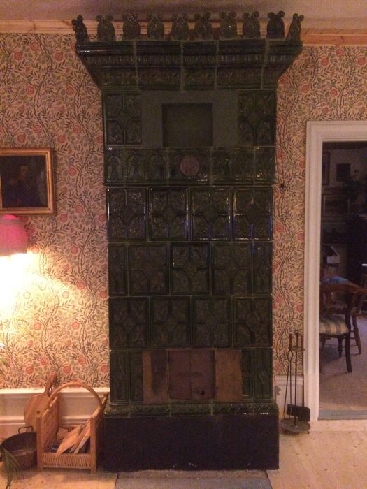 En gammal grön kakelugn i ett rum med mönstrad tapet, vedkorg och eldredskap bredvid, kallad "matsalen".