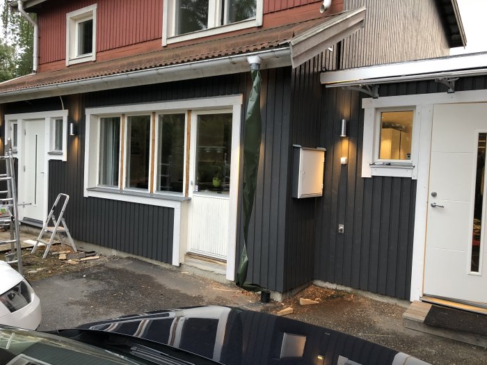 Delvis renoverat tvåvåningshus med nygråmålad nedervåning, vitmålade fönsterkarmar och dörr, samt osläckt stuprör.