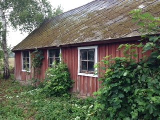 Gammalt lerstenshus med röd träfasad och slitet eternittak omgivet av grönska.