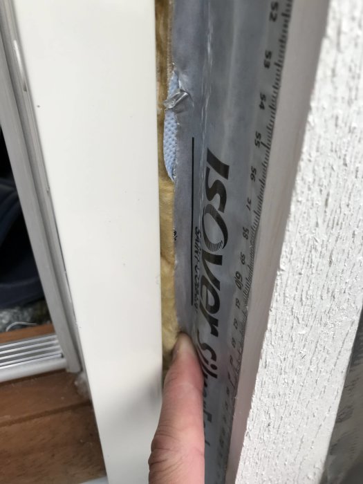 Närbild på en vit altandörr där isolering och en bit av en vindspärrstejp syns vid sidan, med ett finger som pekar på tejpens skarv.