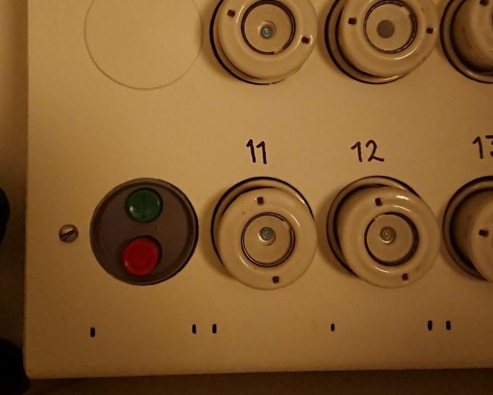 Röd och grön knapp på elcentral omgiven av säkringsdosor märkta 11, 12 och 13.