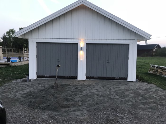 Vit garagebyggnad med två gråa portar och tända utomhuslampor, grushög och skyffel i förgrunden.