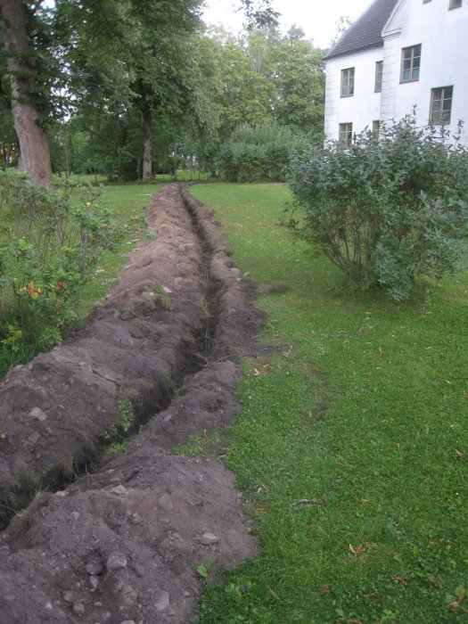 Ett nyligen grävt dike i en gräsmatta, tänkt för fiberkabel, med en vit husfasad i bakgrunden.