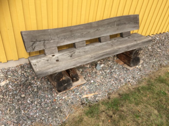 Sliten timmerbänk framför gult hus med brutet ben av två stockar.