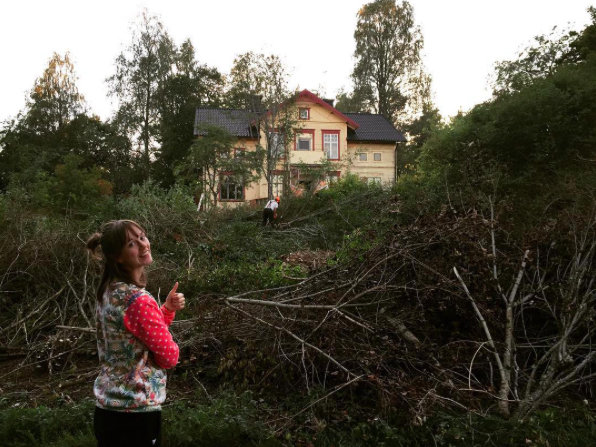 Kvinna ger tummen upp framför ett kraftigt röjt område med grenhögar och ett hus i bakgrunden.