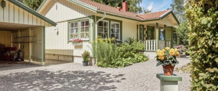 Bild av en charmig villa med beigea fasader och gröna knutar, omgiven av en välskött trädgård och grusgång.