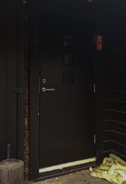 Nyinstallerad svart ytterdörr med fönster och dörrhandtag, i ett hus med mörka träväggar.