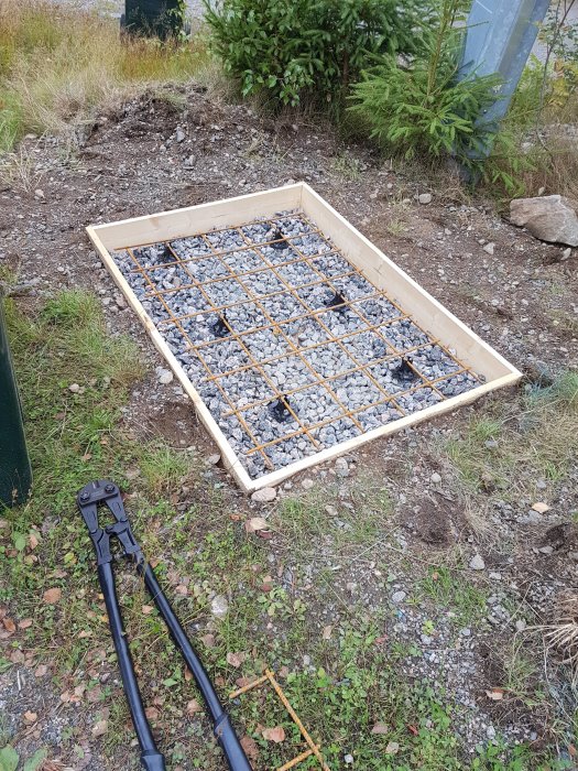 En liten form för betonggjutning av en platta klargjord med armeringsjärn och grus, placerad på en ojämn gräs- och grusmark.