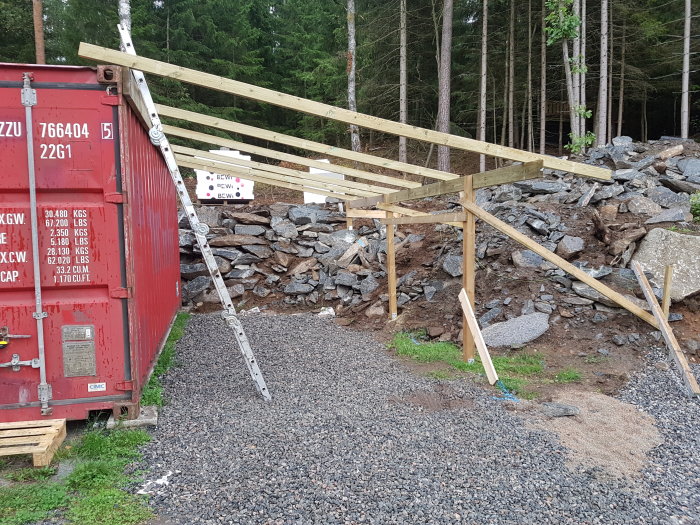 Pågående bygge av ett takstomme i trä med en stege lutad mot en röd container, plats för överbliven betong synlig.