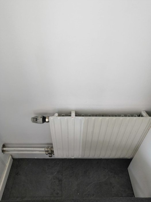 Radiator med anslutna rör och termostatventil mot en vit vägg ovanför ett mörkt golv.