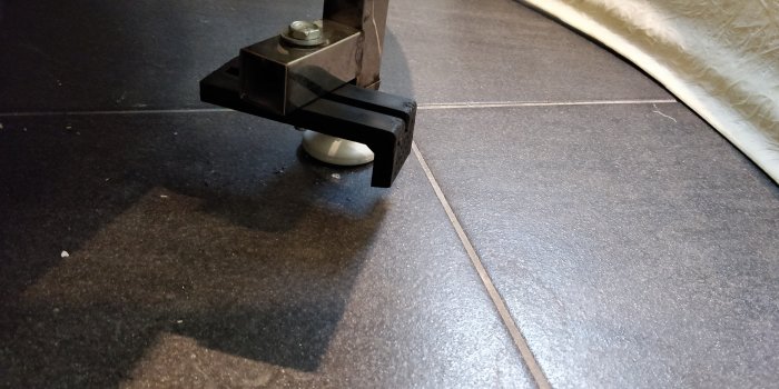 Svart L-format badkarsben ställt in på en justerbar fot, placerad på ett mörkt golv med en del av fronten synlig.