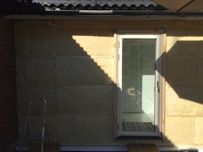 Dörr med glas insatt i en isolerad vägg med vindskydd och synliga reglar, oavslutad fasadrenovering.