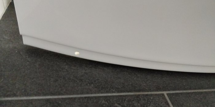 Nedre delen av en skåpfront med ett synligt skruvhål mot en grå klinkergolv.