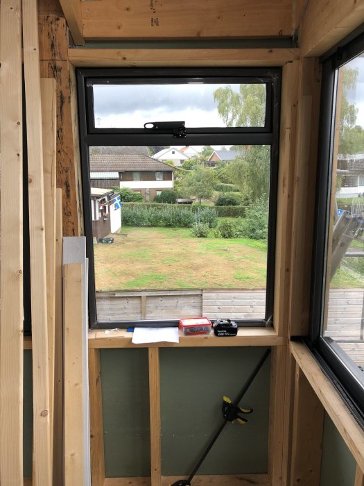 Utsikt från ett ofärdigt rum under renovering med synliga trästommar och ett öppet fönster som visar en trädgård och hus.