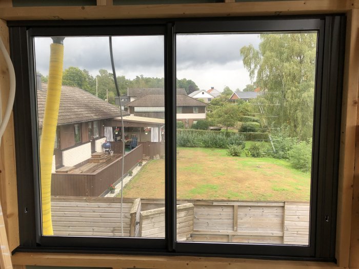 Utsikt från ett fönster över en trädgård med gräsmatta, träd och en träterrass.