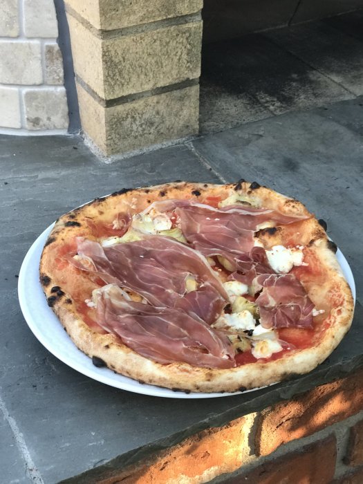 Hembakad pizza i napolitansk stil med tomatsås, mozzarella och prosciutto på en tallrik framför en stenugn.