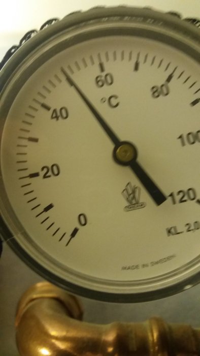 Analog termometer som visar 55 grader Celsius, placerad över en mässingsrörledning.