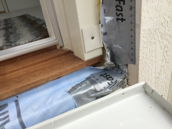 Avlägsnade aluminiumprofiler vid en dörrkarm som avslöjar en vindduk och mätsticka visar 15 mm springa.