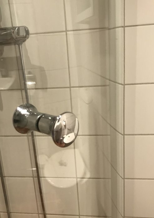 Kromat duschväggsgångjärn med avbruten grå plastdel på kakelvägg.