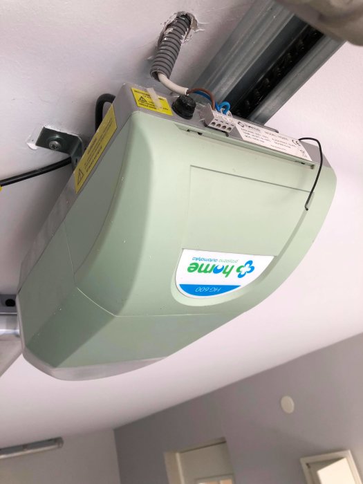 Motordriven garageportsöppnare med synliga kablar och varningsetiketter monterad i taket.