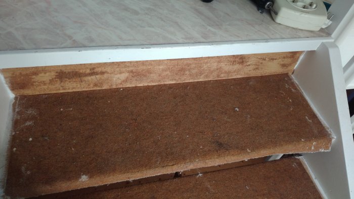 Delvis vitmålad trappa med sliten brun filtmatta och trädetaljer där färg släppt och träådring syns.