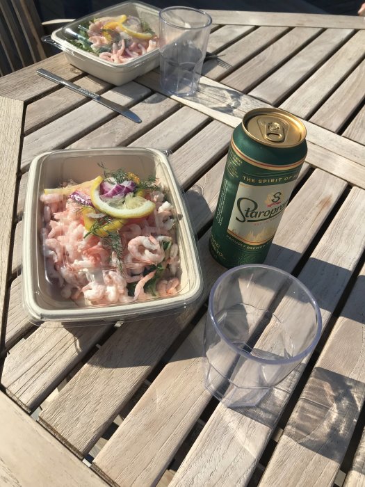 Måltid på träbord med räksallad, ölburk och tomt glas, soligt utomhus.