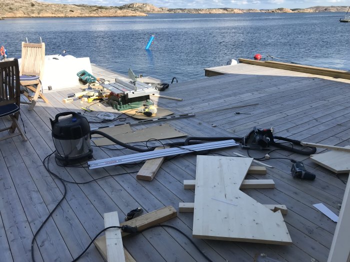 Byggmaterial och verktyg utspridda på en trädäck vid sjön under pågående arbete med en spiraltrappa.
