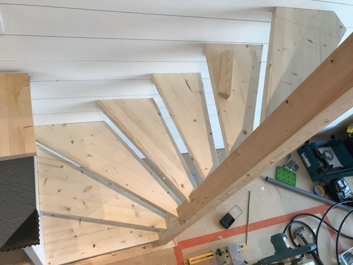 Delvis färdigställd spiraltrappa i trä under konstruktion, med verktyg och byggmaterial synliga.