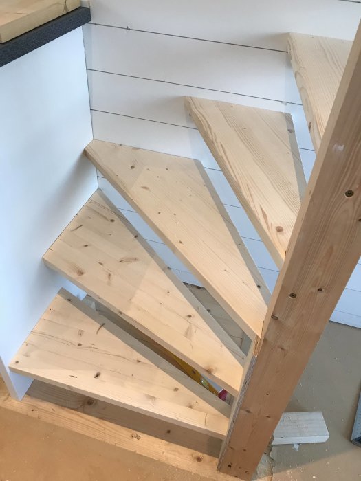 Nybyggd träspiraltrappa under konstruktion i ett ljust rum, delvis uppritad på limfogen, nära vägg.