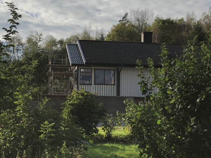 Ett hus med nylagt svart tak och ställningar kring altan i en lummig trädgård.