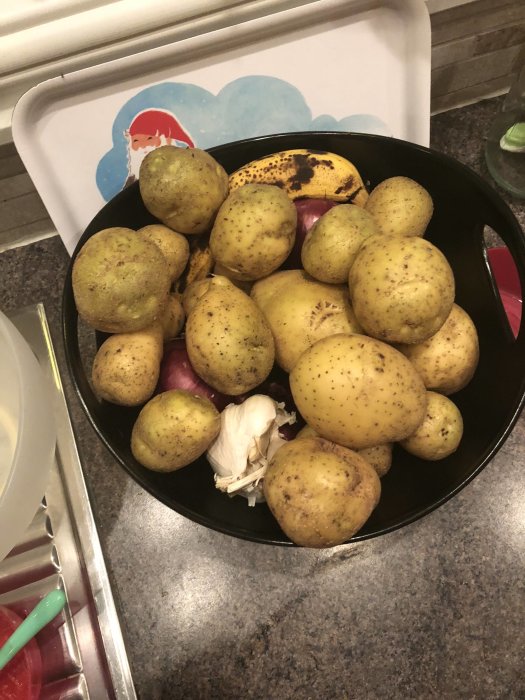 Skördad potatis och vitlök i en svart skål på ett köksbänk, med en del mognad banan och rödlök.