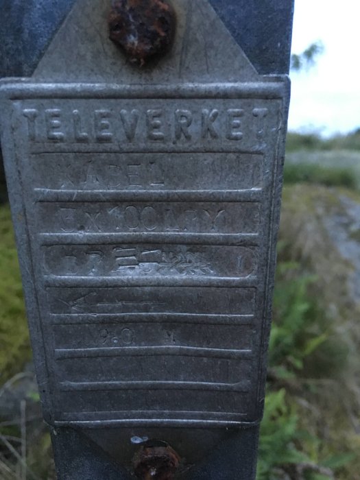 Närbild på en skylt med texten "Televerket" och teknisk information om en kabel, rostig och monterad utomhus.