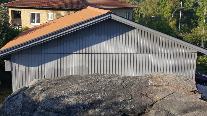 Nymålat grått garage med tegelröd takbeklädnad nära stor sten och grönska.