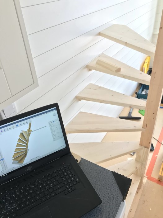 Ofärdig trätrappa med tre steg kvar att bygga, bredvid en laptop som visar ritningar av trappan.