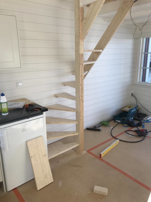 Pågående bygge av trätrappa med tre osatta steg, verktyg och material utspridda i ett ljustrum.