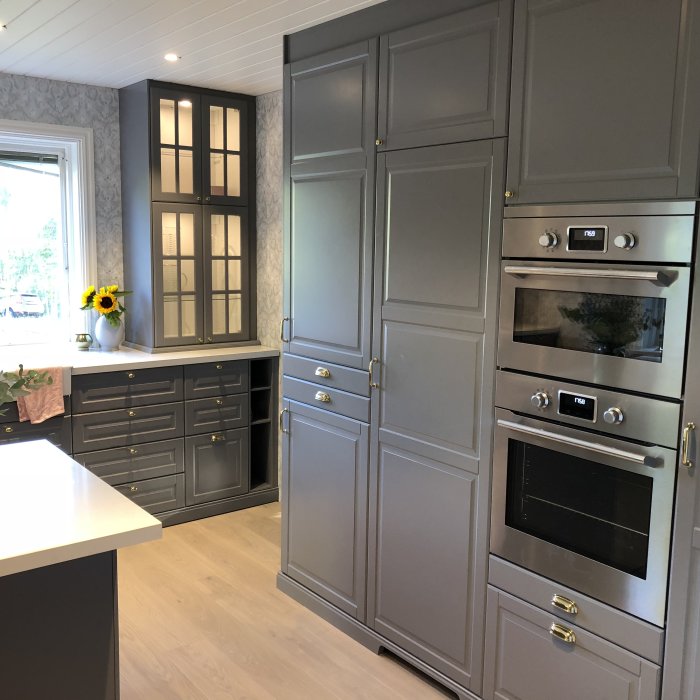 Modernt kök med grå skåp, inbyggda ugnar och en vit bänkskiva, dekorerat med solrosor.