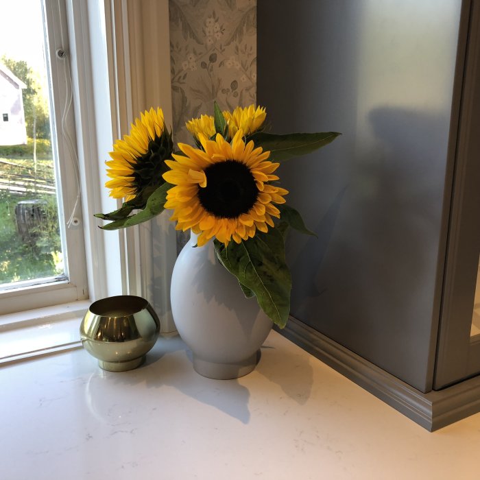 Vas med solrosor på fönsterbräda bredvid en speglad yta och en liten grön vas.