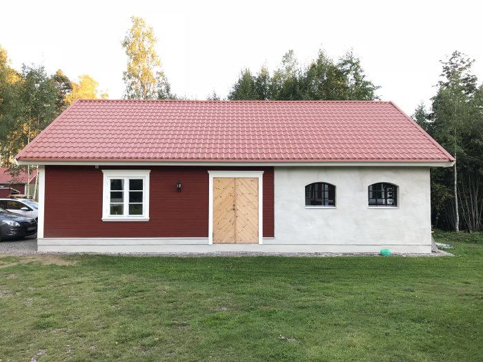 Nästan färdigt garage med röd putsad fasad, obehandlad dörr och vita fönster under tak med röda tegelpannor.