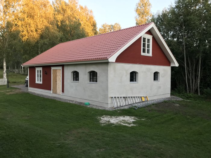 Nybyggt garage med rött tak och vit putsad fasad, fönster på plats, omålad dörr och saknade hängrännor.