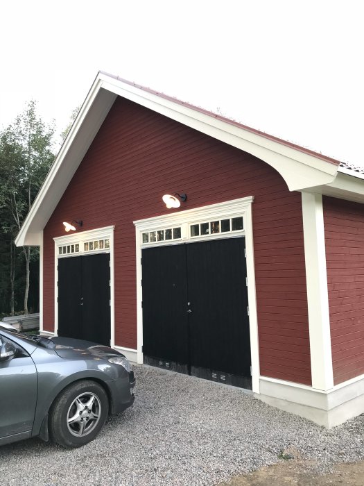 Rödmålat garage med svarta dubbelportar, överljus och 1920-talslampor ovanför, i dagsljus.