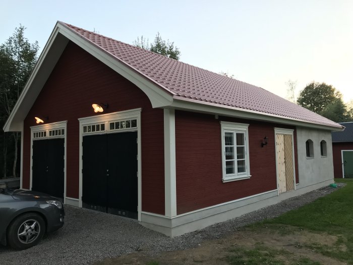 Nästan färdigt rött garage med svarta portar, vita fönster och ommålad dörr samt 20-tals lampor ovanför portarna.