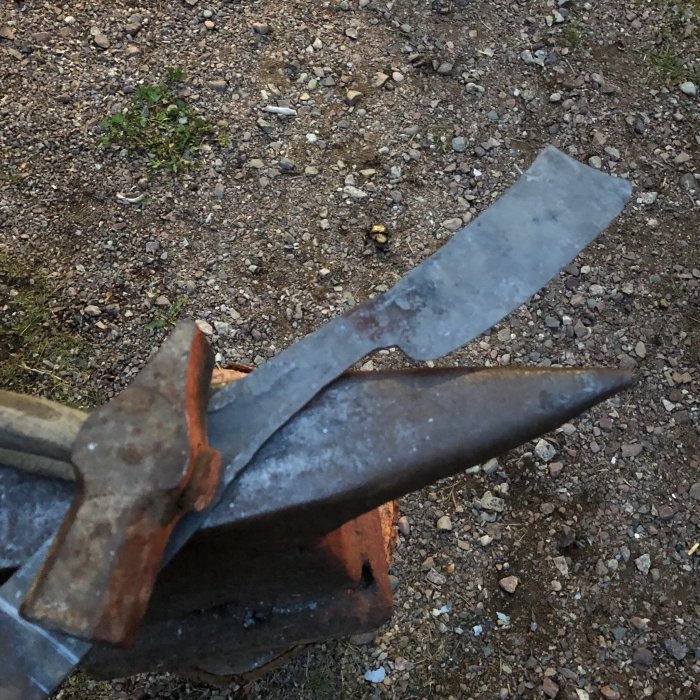 Handsmidda metallföremål i ett skruvstycke med grusig mark i bakgrunden.
