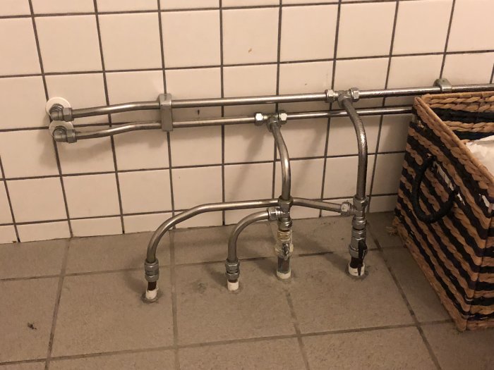 Oorganiserade rörledningar och en flätad korg i ett klubbhus badrum.