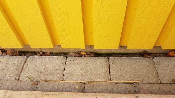 Trottoarstenar lagda intill en gul husvägg med ett utrymme på några centimeter till husets grund.