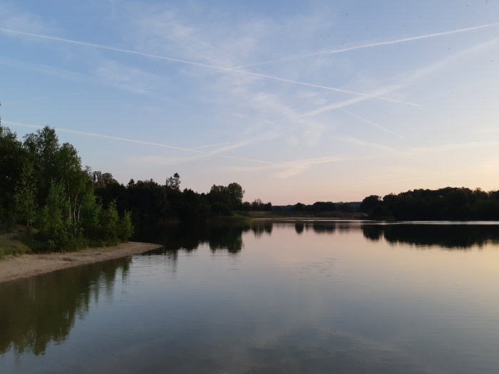 En lugn sjö vid skymningen med träd och klar himmel med flygplansstrimmor.